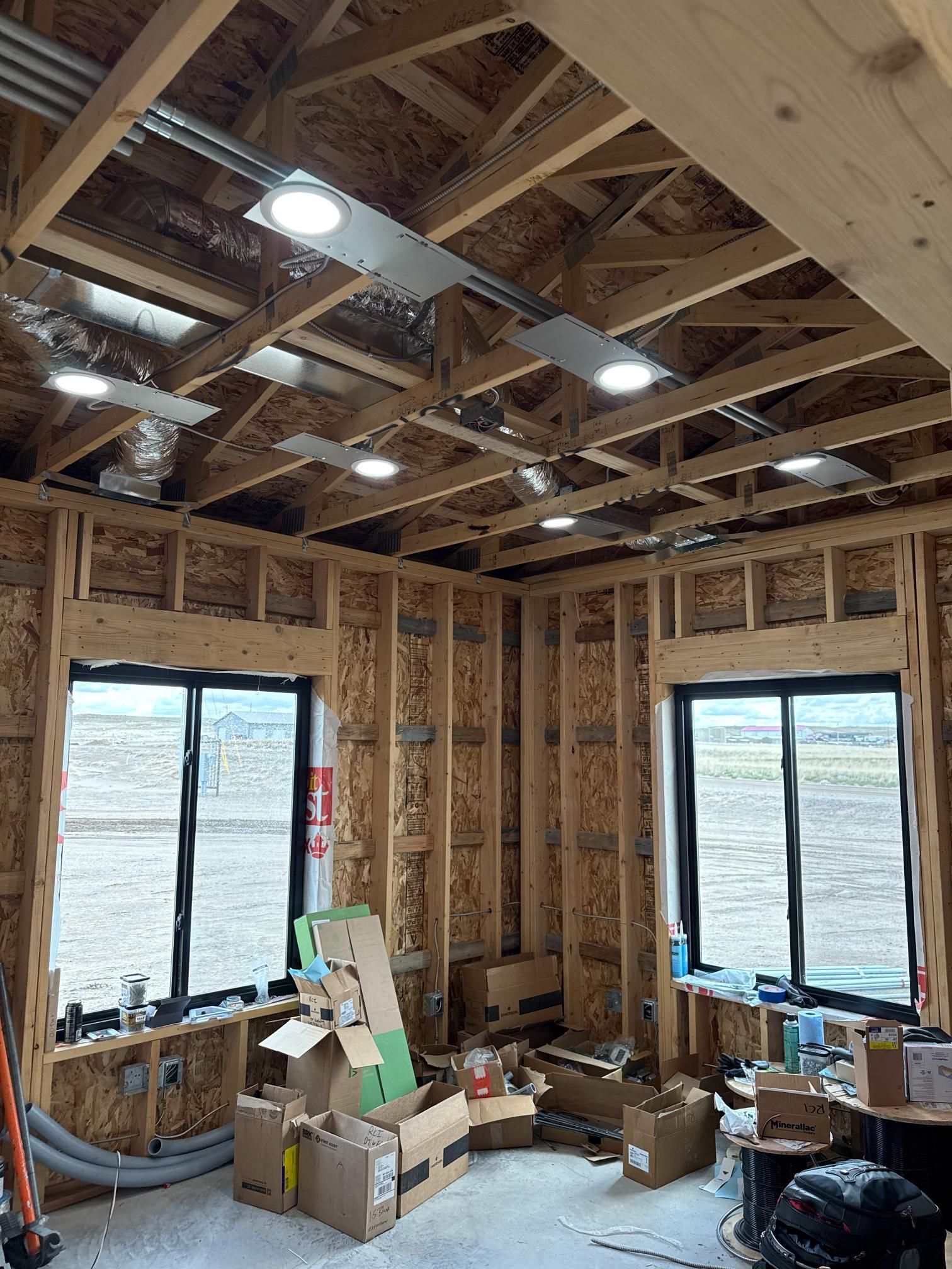 Interior of a room under construction with exposed wooden framing, windows, and overhead lighting fixtures.