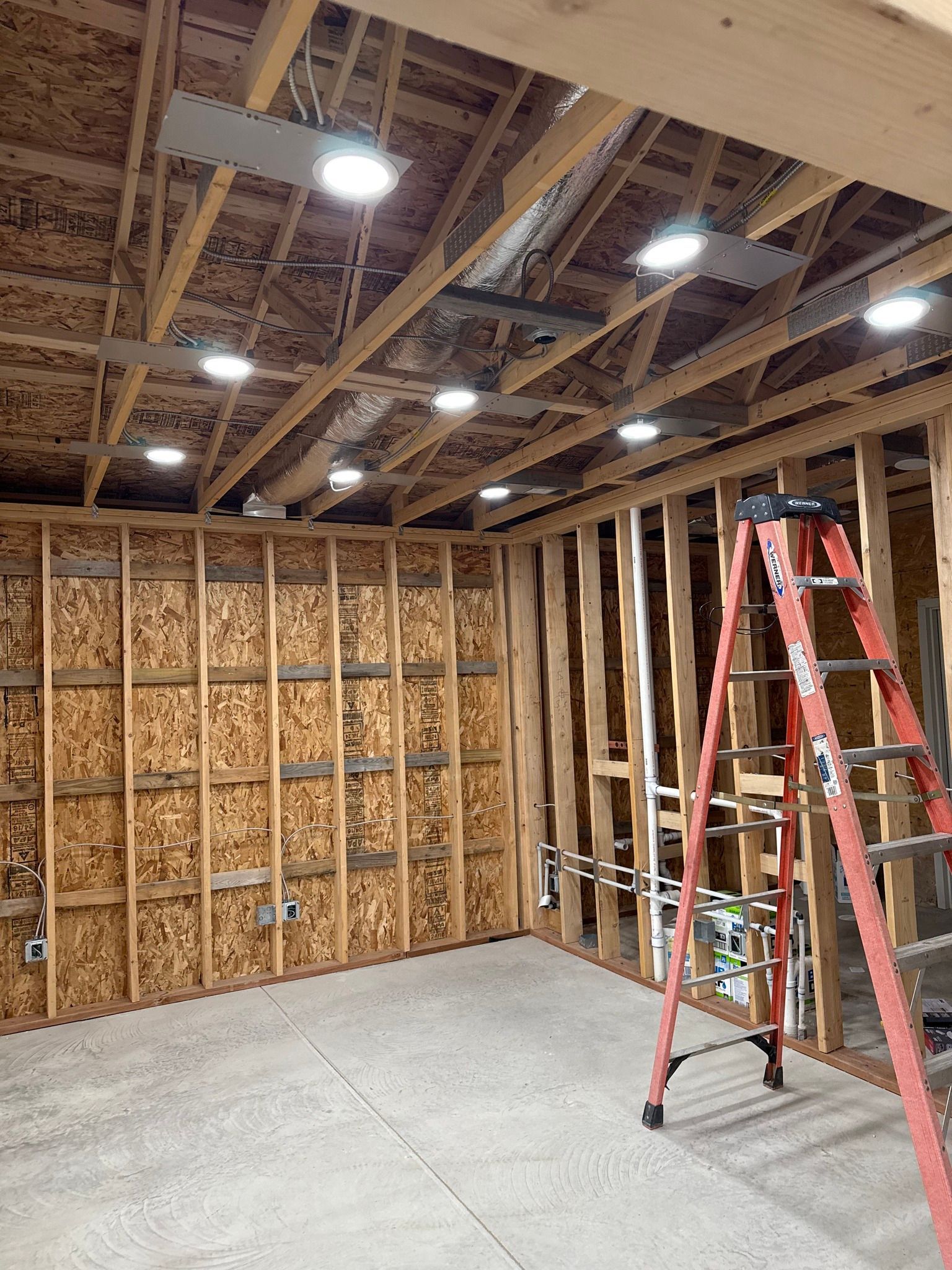 Interior of a room under construction with exposed wooden framing, recessed lights, and a red ladder.