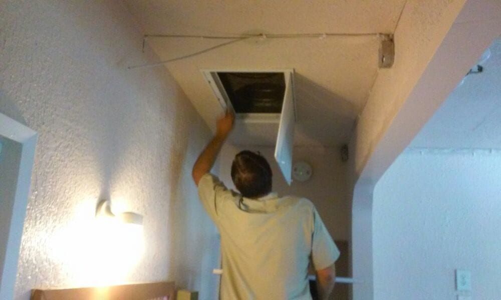 Man opening attic access panel in a hallway. White ceiling, beige walls, overhead light fixtures.