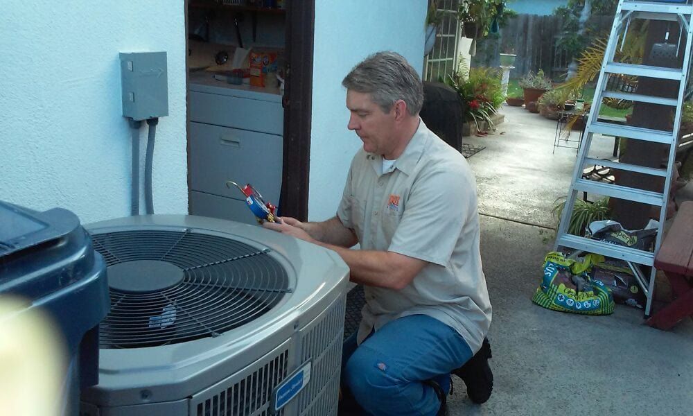 A man kneels by an AC unit, examining it with a tool. Outdoor setting, white building, ladder, and plants.