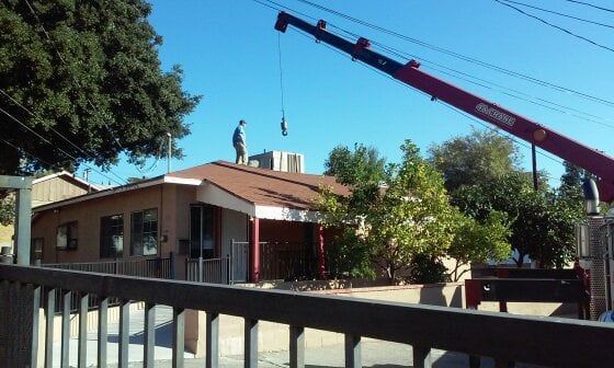 A crane lifts something from a house roof. A person stands on the roof. Bright blue sky.