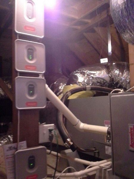 Four gray air quality sensors mounted on a wooden beam in an attic. Wires and HVAC equipment visible.