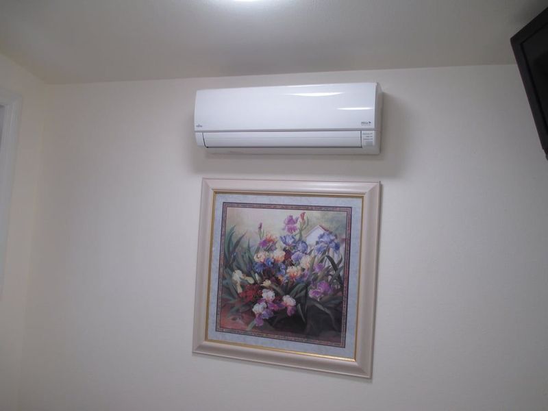 White wall-mounted air conditioner above a framed floral painting on a light-colored wall.
