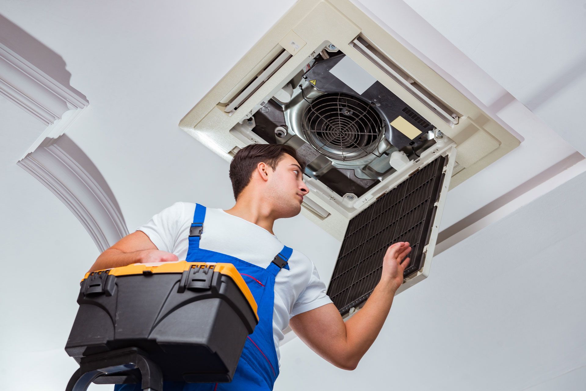 HVAC technician on a ladder inspects a ceiling-mounted air conditioning unit, holding a filter and toolbox.