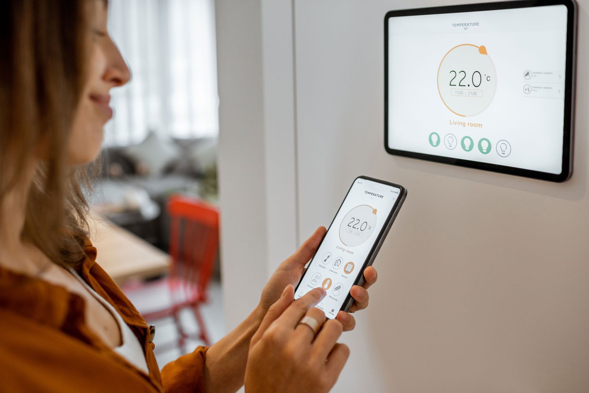 Woman using smartphone to control a smart thermostat on a wall, displaying the temperature.