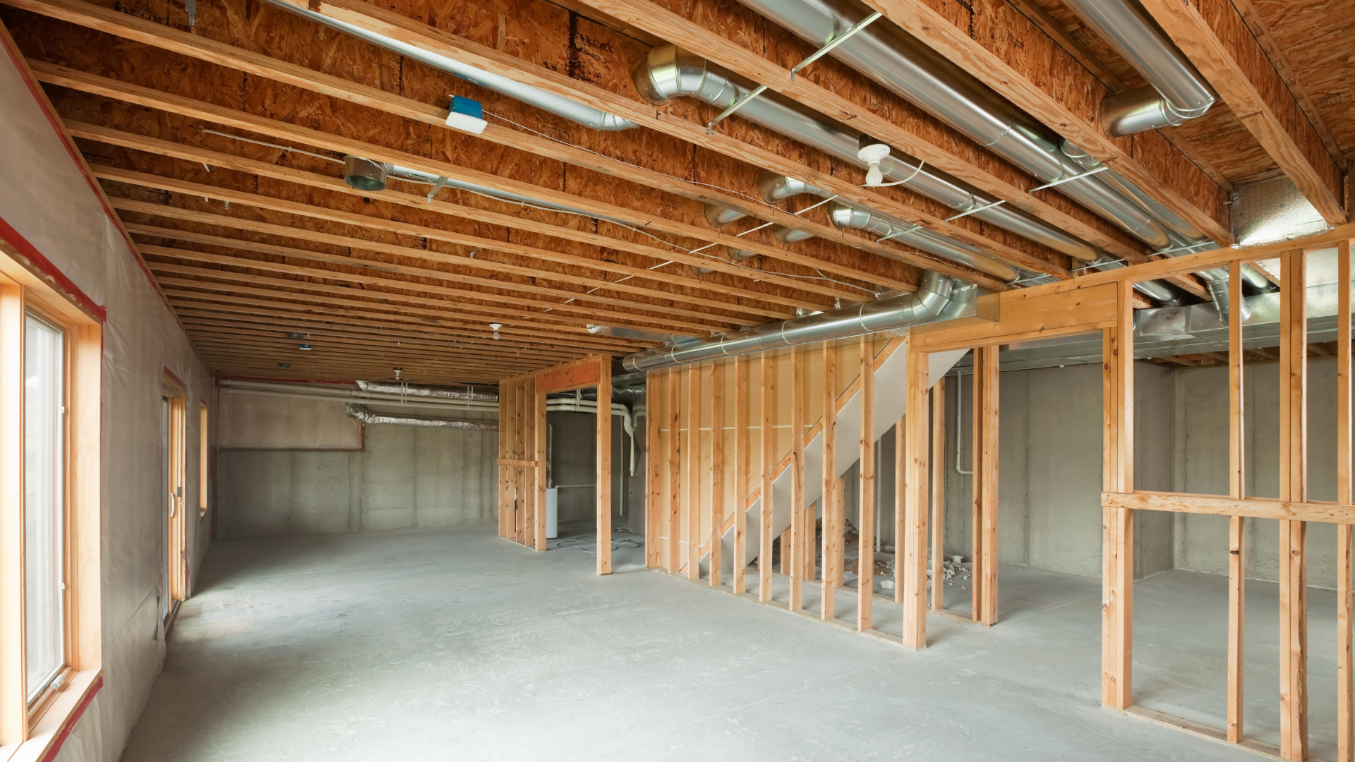 Unfinished basement with exposed wooden joists, concrete walls, ductwork, and partially framed interior walls.