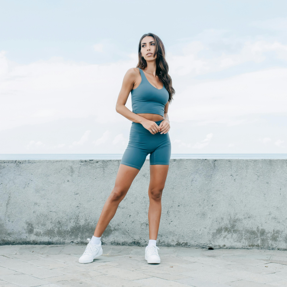A woman in a crop top and shorts is standing in front of a concrete wall.
