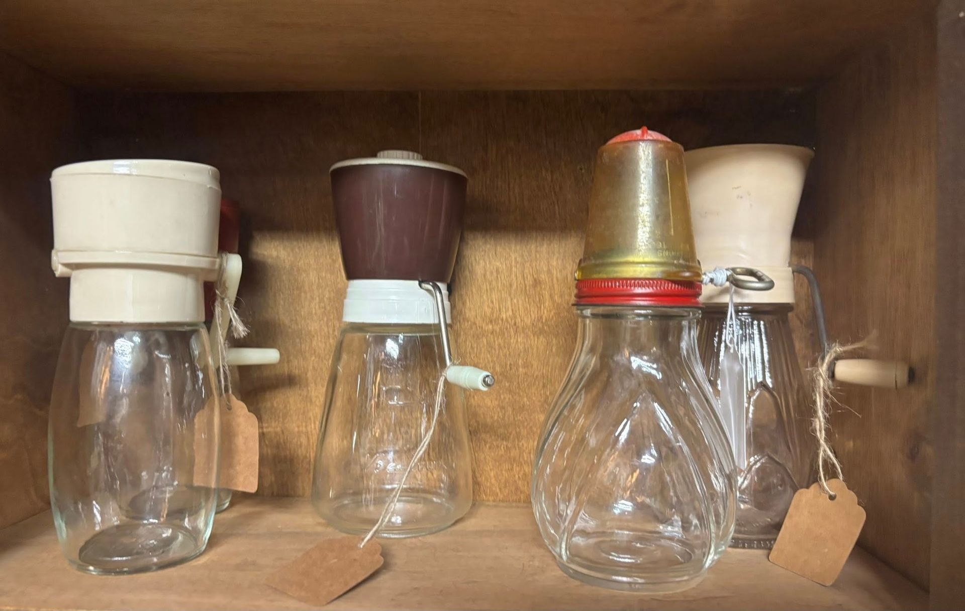 Four vintage glass grinders with various colored tops sit on a wooden shelf.