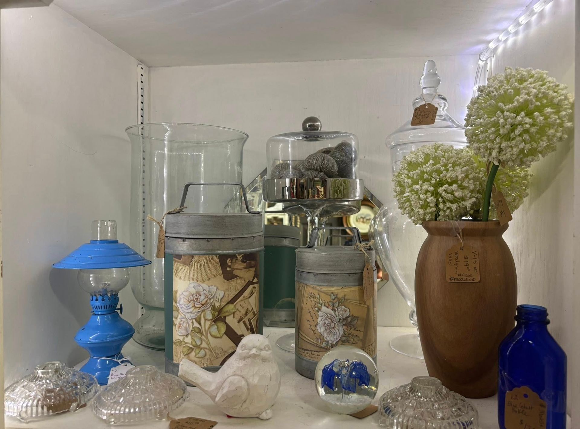 Shelf display with glass jars, a blue lamp, and decorative items, including a vase with flowers.