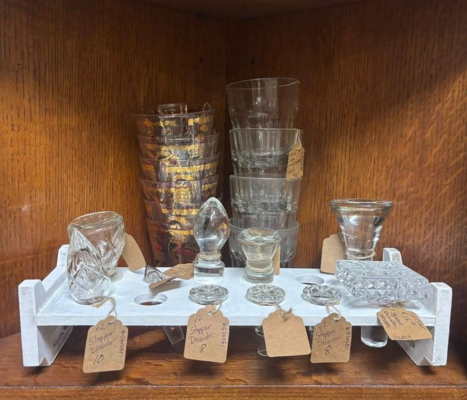 Various antique glassware for sale on a white shelf inside a wooden cabinet.