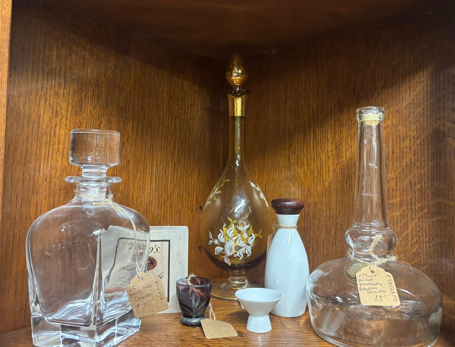 Bottles and small items on a wooden shelf. Includes clear, amber and white glass. Tags attached.