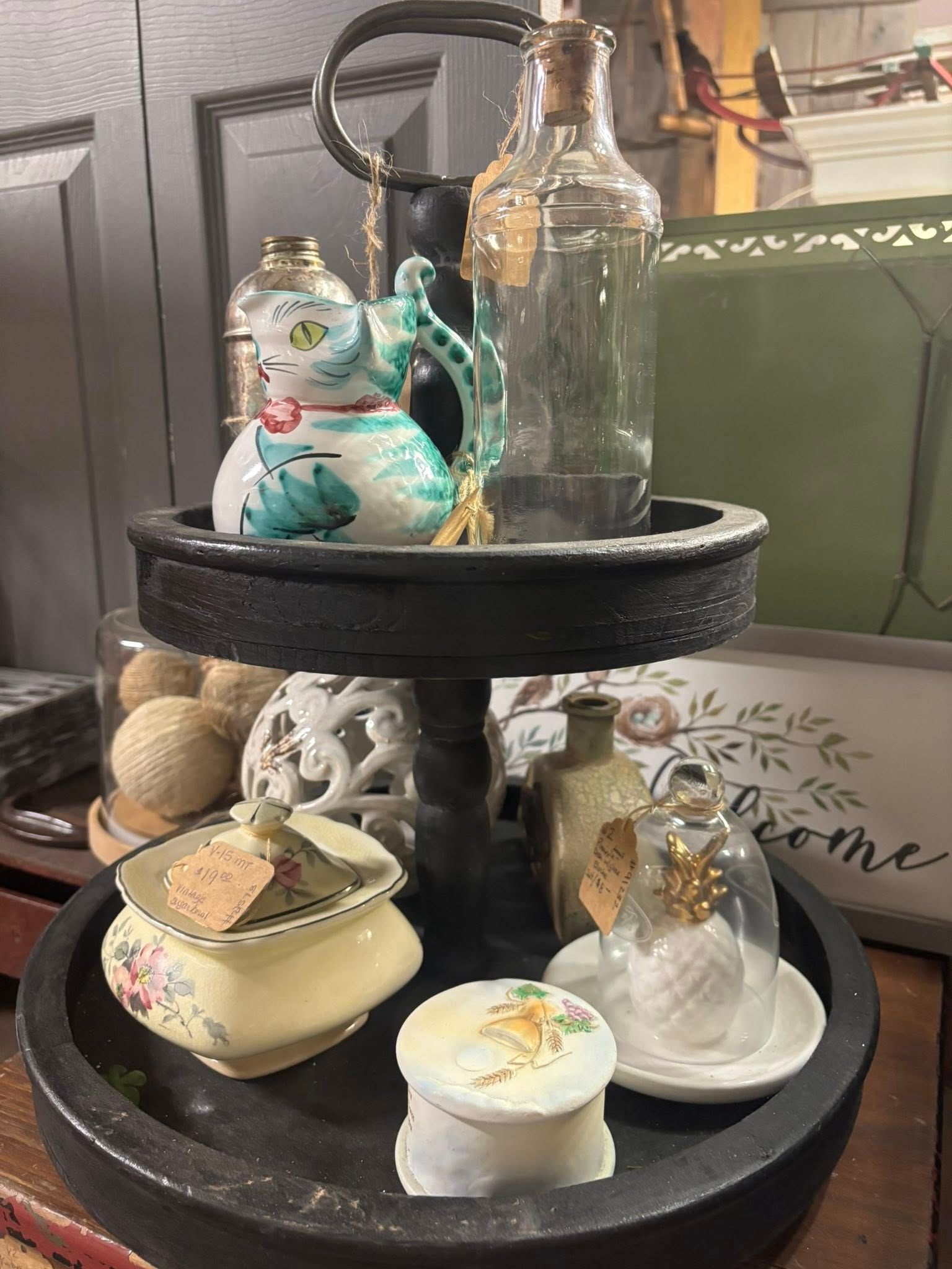 Two-tiered black tray with decorative items: cat creamer, bottles, trinket boxes, and a pineapple under glass.