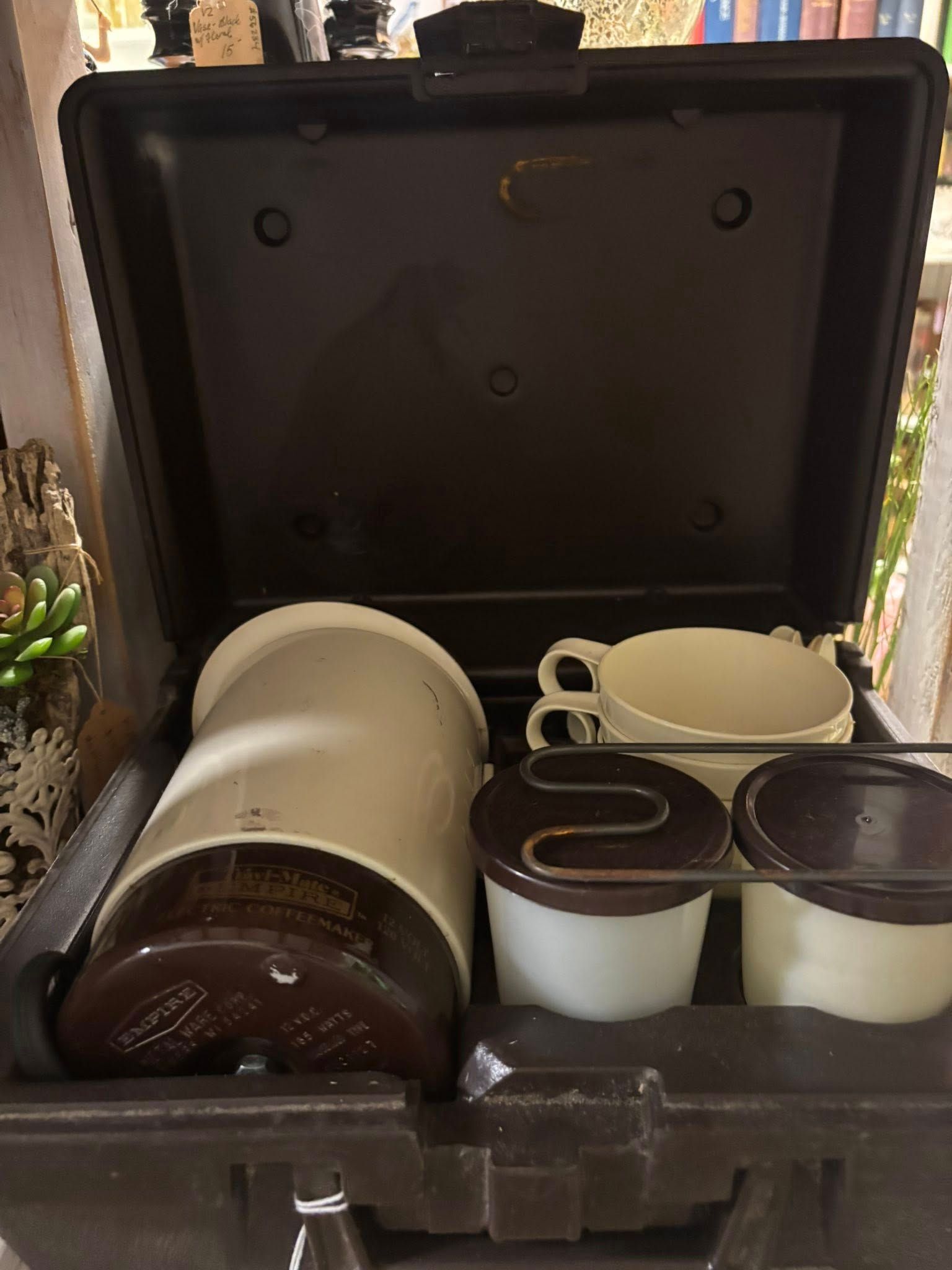 A black case holds a cream-colored coffee maker, two cups, and two small containers; a brown lid is a detail.
