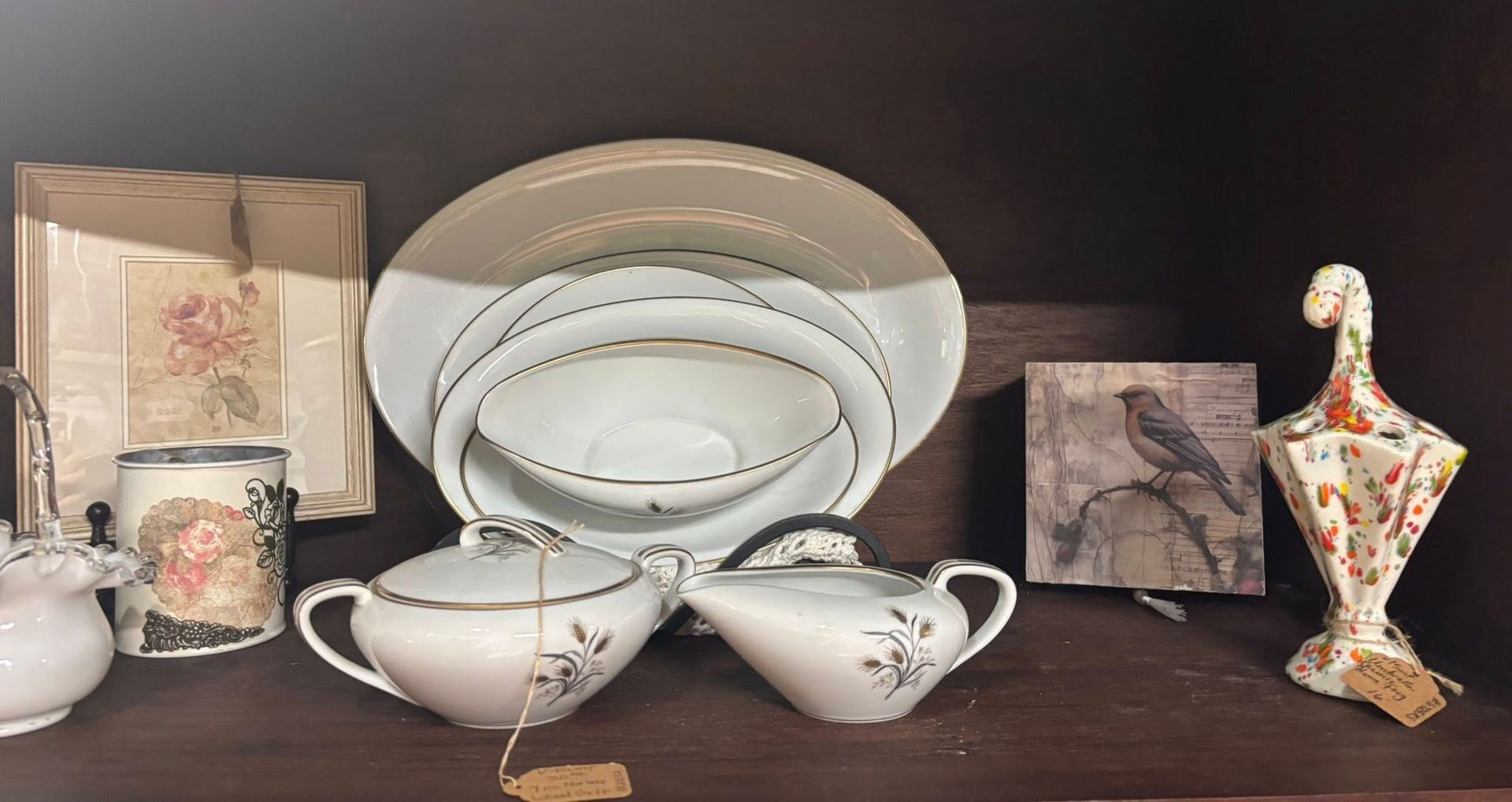 China and decorative objects on a dark shelf. White and gold dishes, framed artwork, and a ceramic bird.