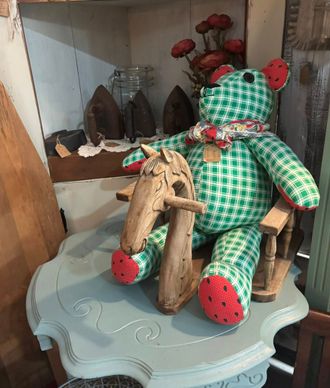 Green gingham bear in rocking chair, holding wooden horse on a light blue table in a shop.
