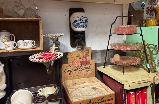 Various antique glassware for sale on a white shelf inside a wooden cabinet.