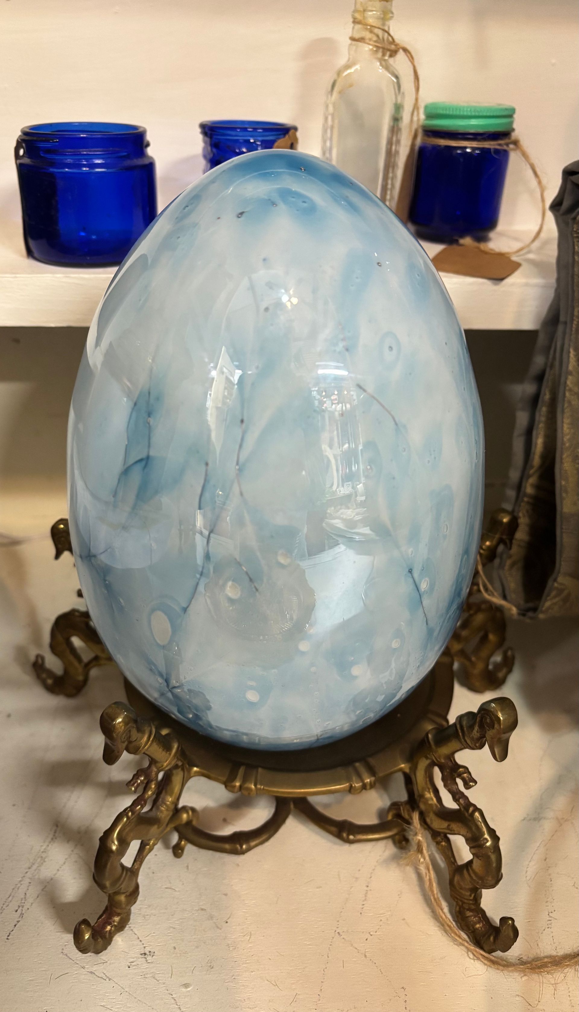 Large blue and white marbled egg on ornate brass stand; jars in background
.