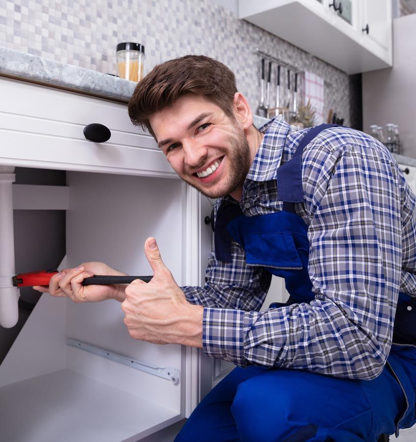 A plumber is fixing a sink in a kitchen and giving a thumbs up.
