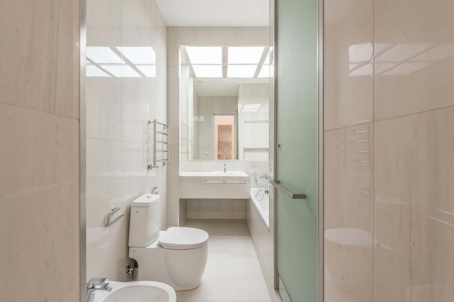 Bright, narrow bathroom with white tiles, toilet, bidet, vanity, and frosted glass shower door.