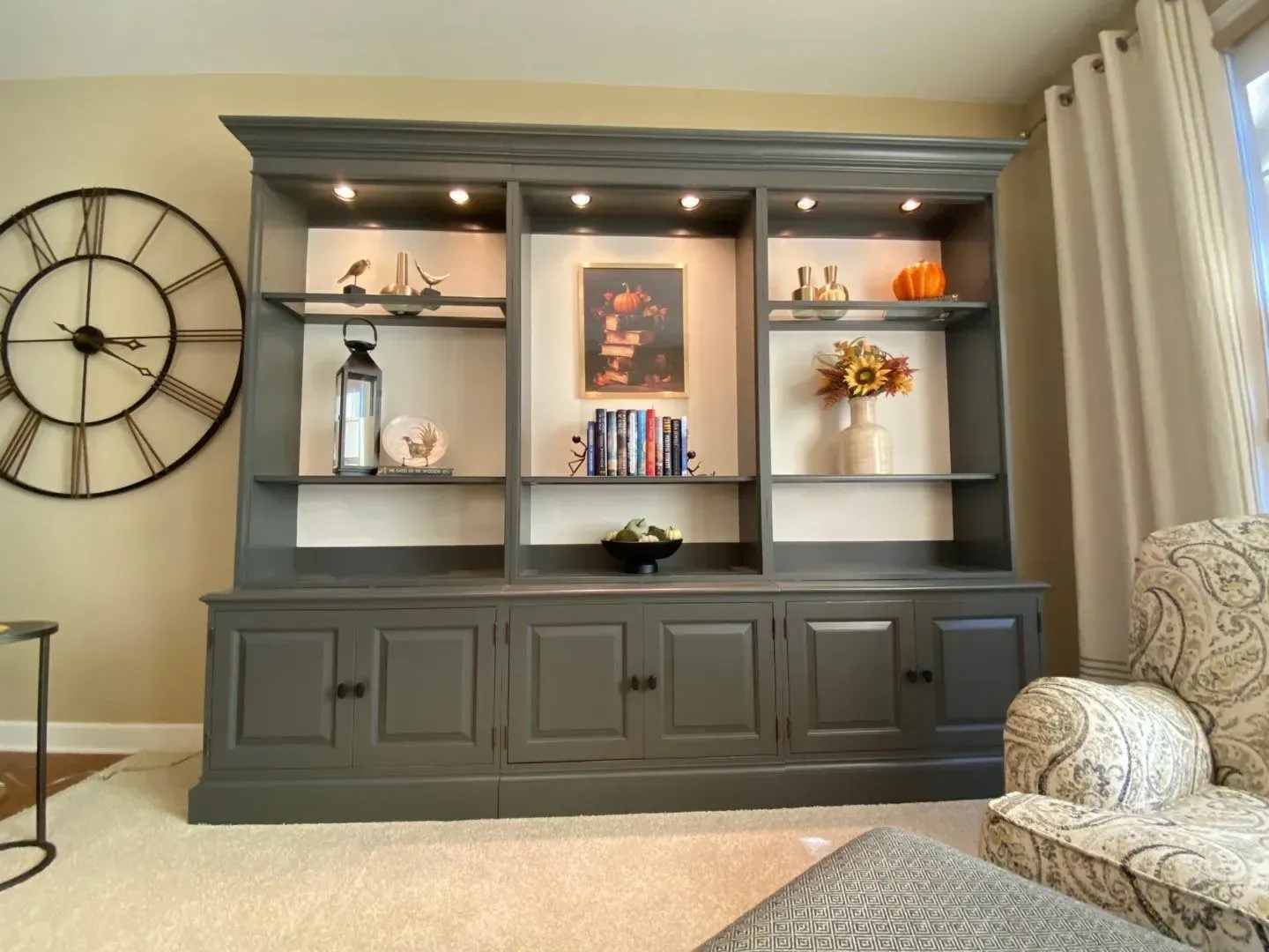 Built-in shelving and cabinet with decor, large wall clock, and armchair in a cozy living room