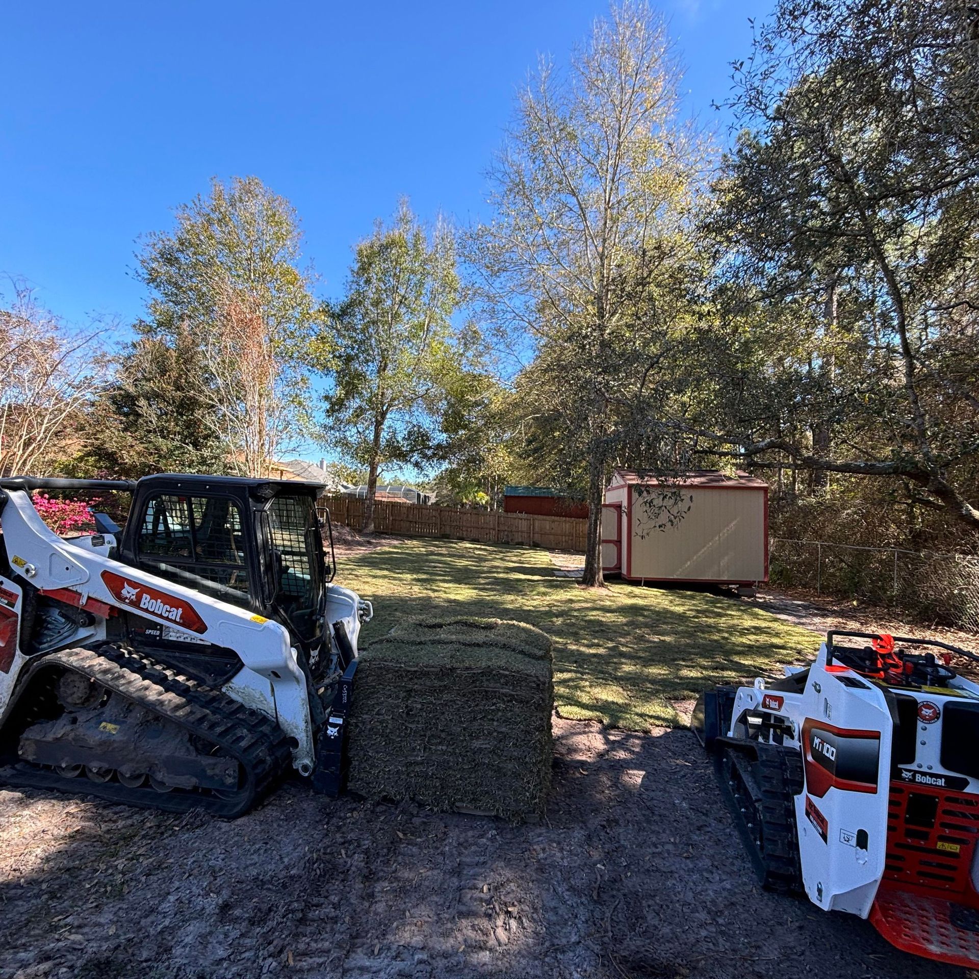Two Bobcat machines flank a square bale of sod in a grassy area with trees and a small shed.