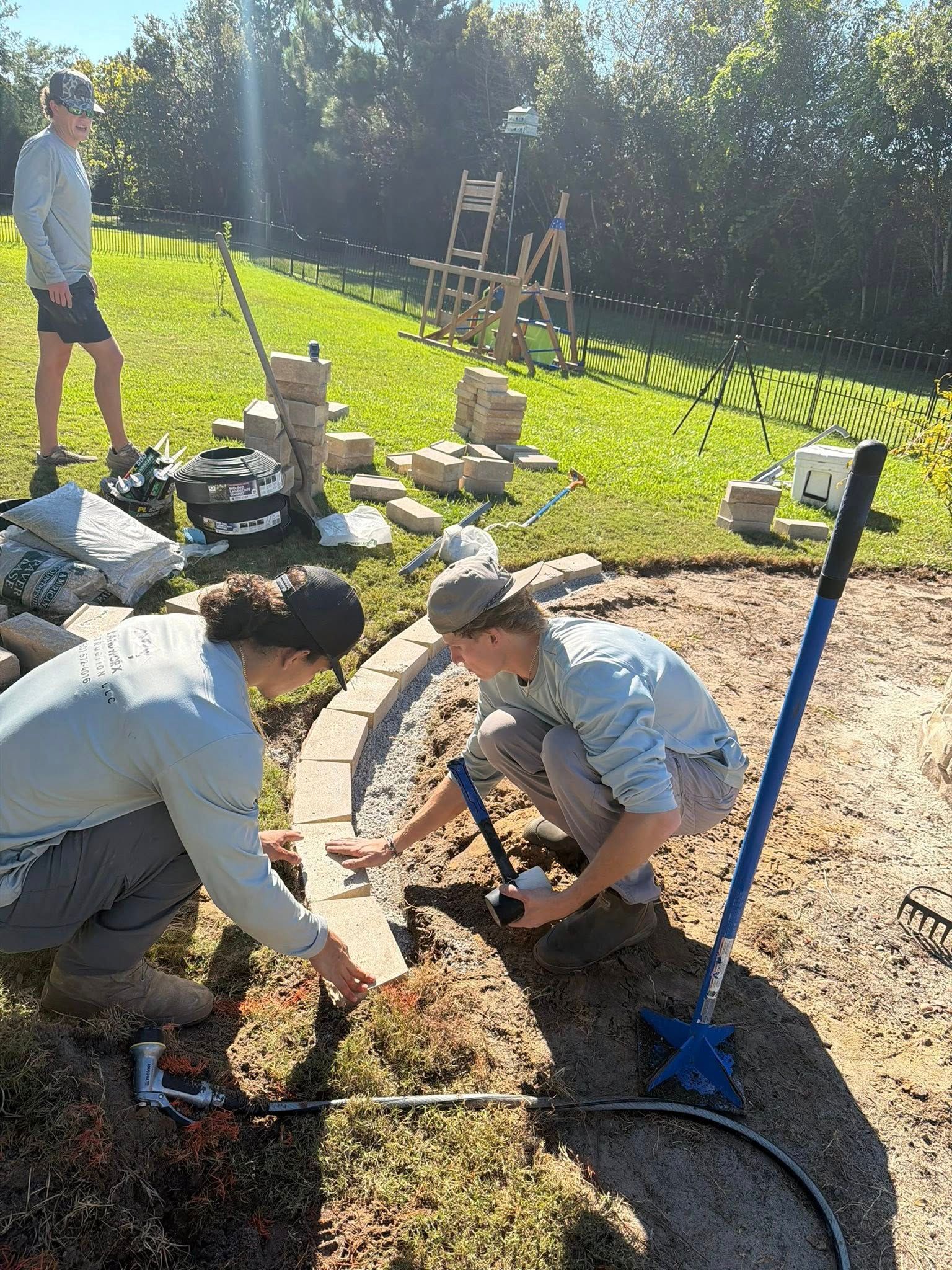 Two people laying bricks around a garden bed in a sunny yard. Another person stands nearby.