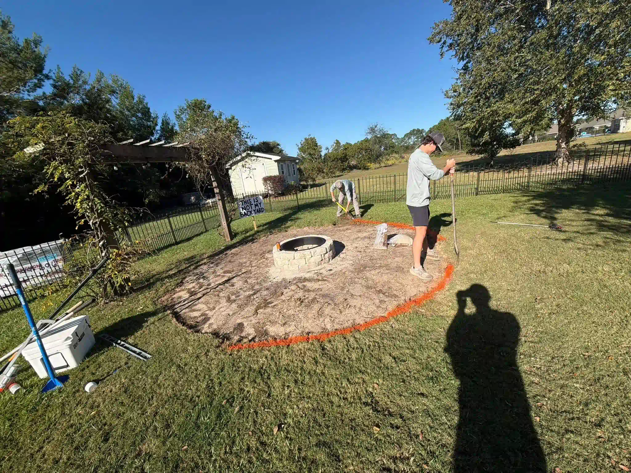Person outlining a circular area in a grassy yard, with a fire pit in the center. Bright sunny day.