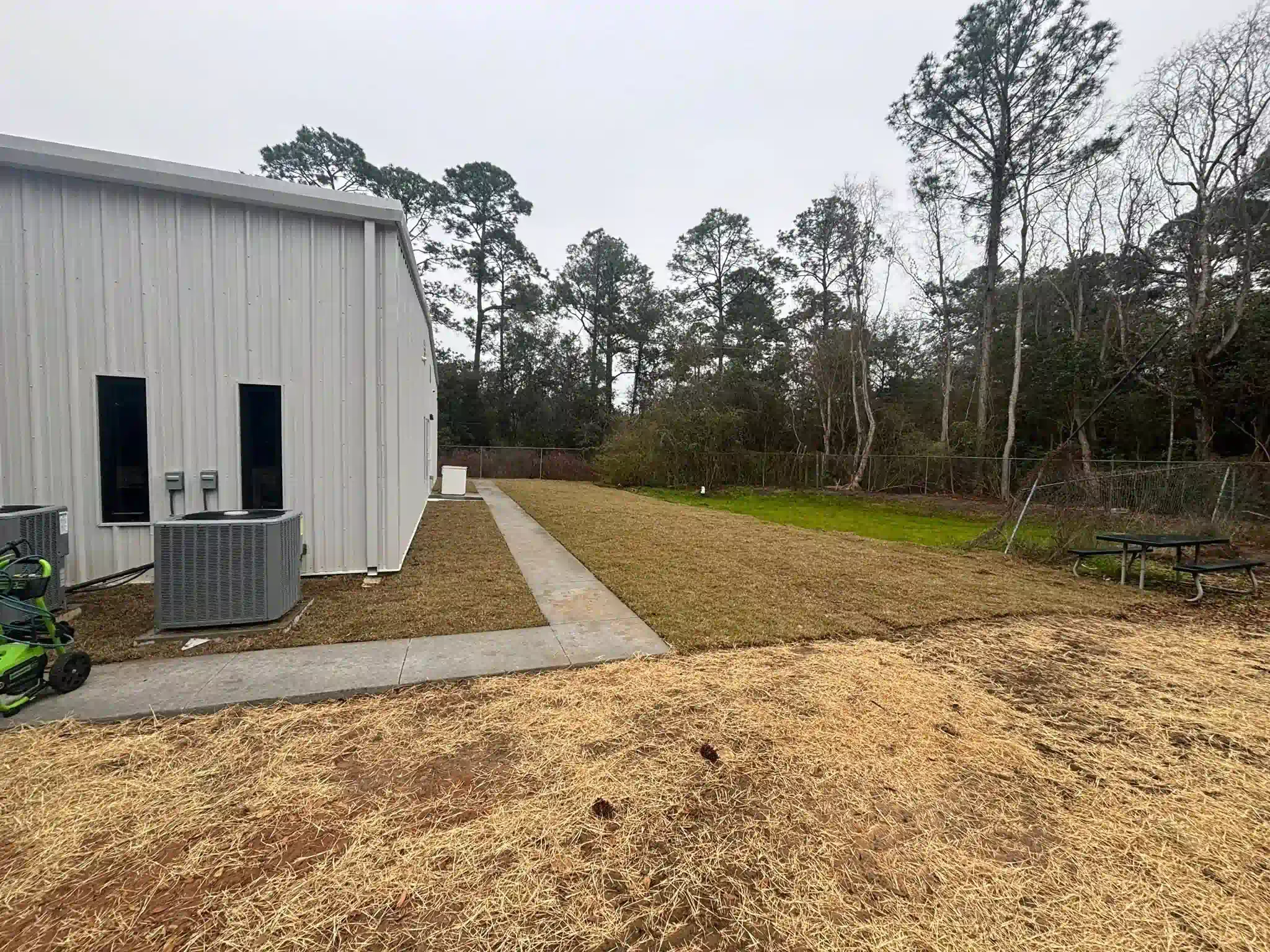 A metal-sided building with a concrete path leading to a grassy area with trees under an overcast sky.