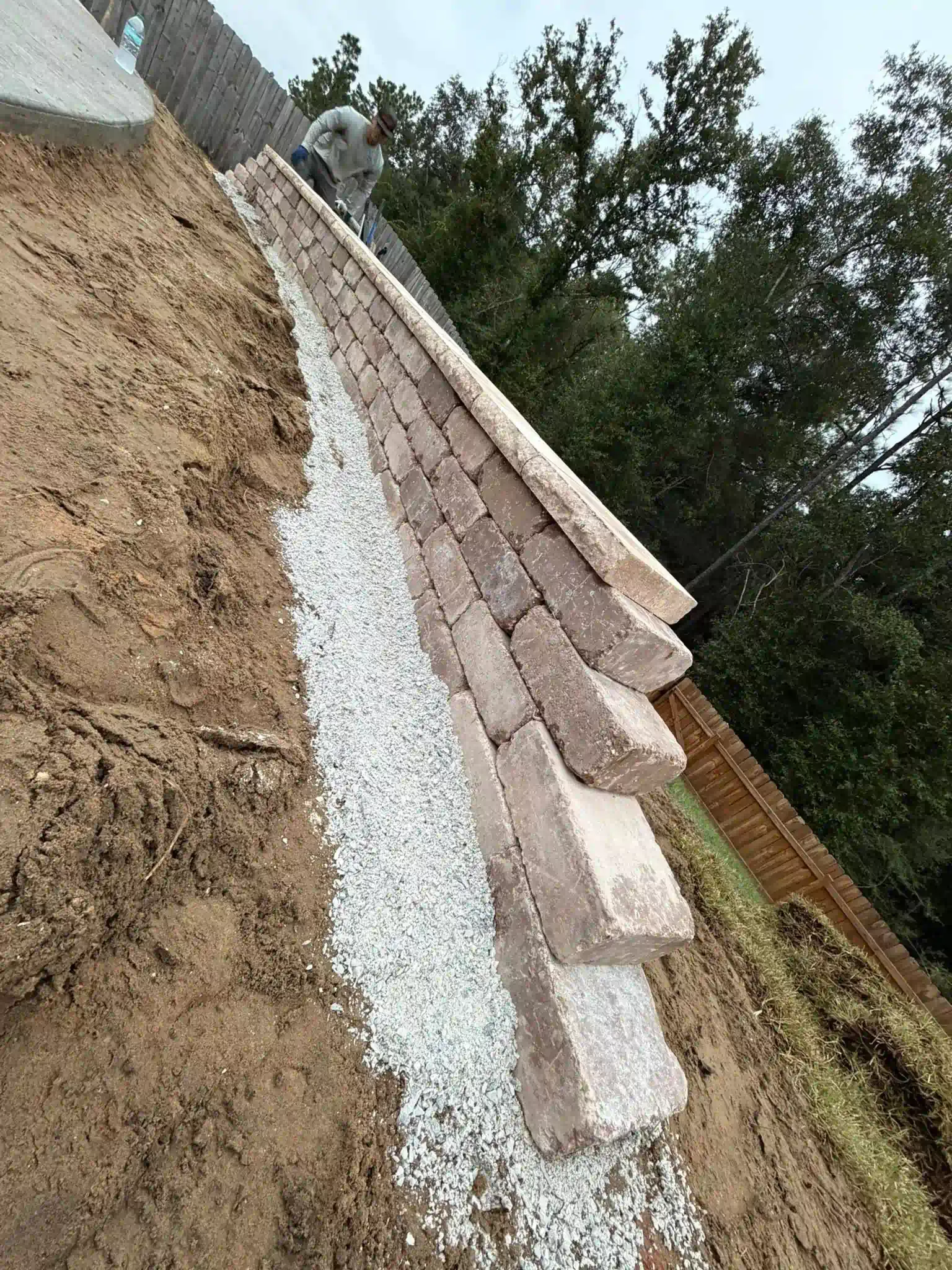 Construction of a retaining wall: blocks, gravel, and dirt on a hillside. A person is working in the background.