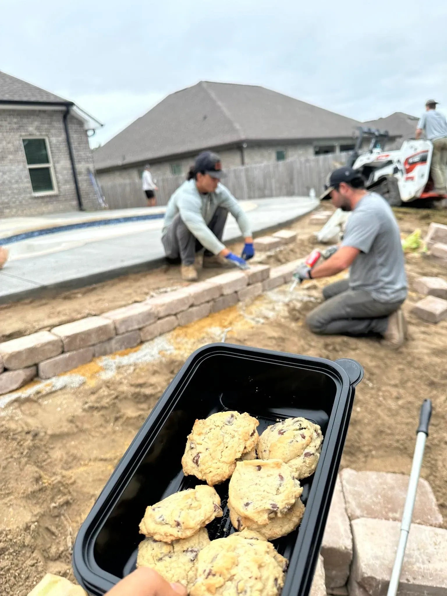 Cookies in foreground; two people working on a brick retaining wall near a pool under construction.
