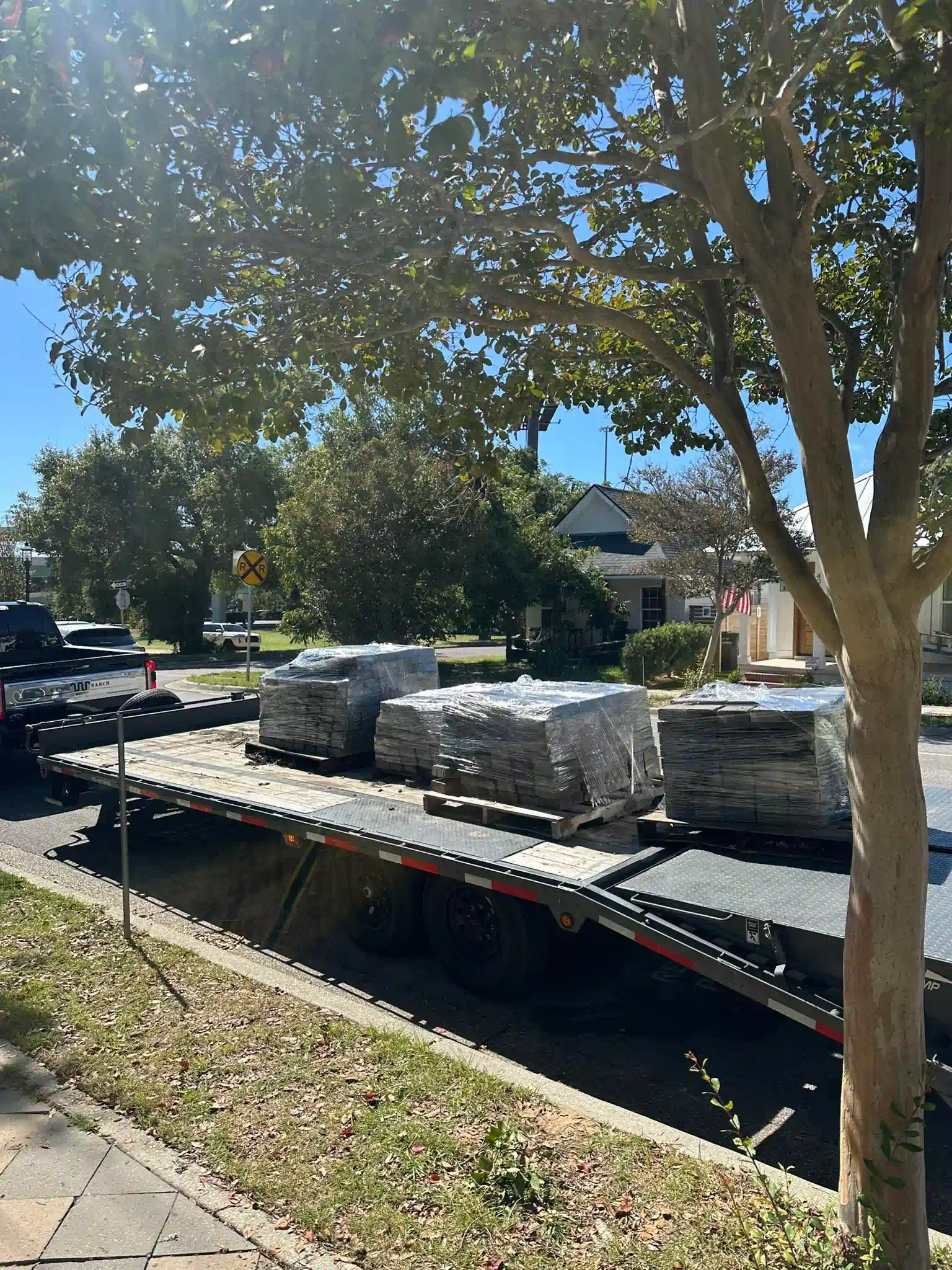 Truck bed loaded with gray stone pavers on a street under a tree on a sunny day.