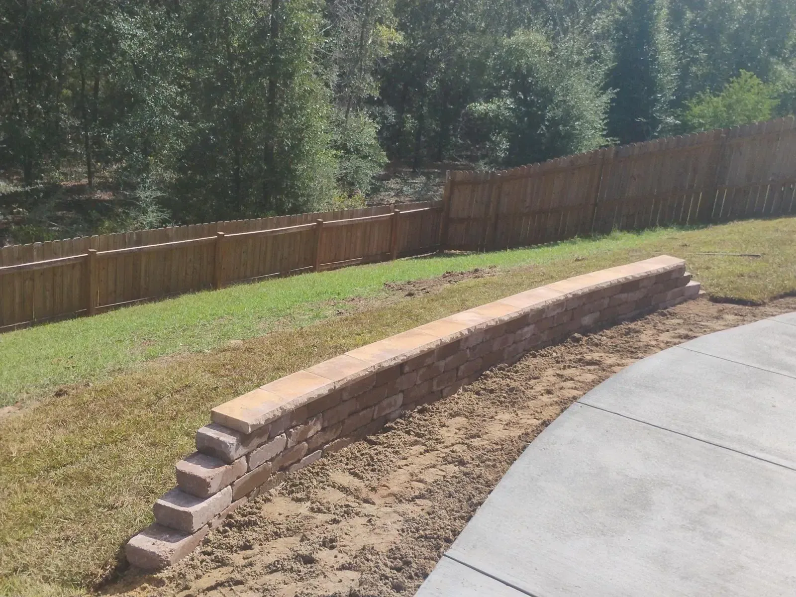 Brick retaining wall on a hillside with a wooden fence and concrete patio.