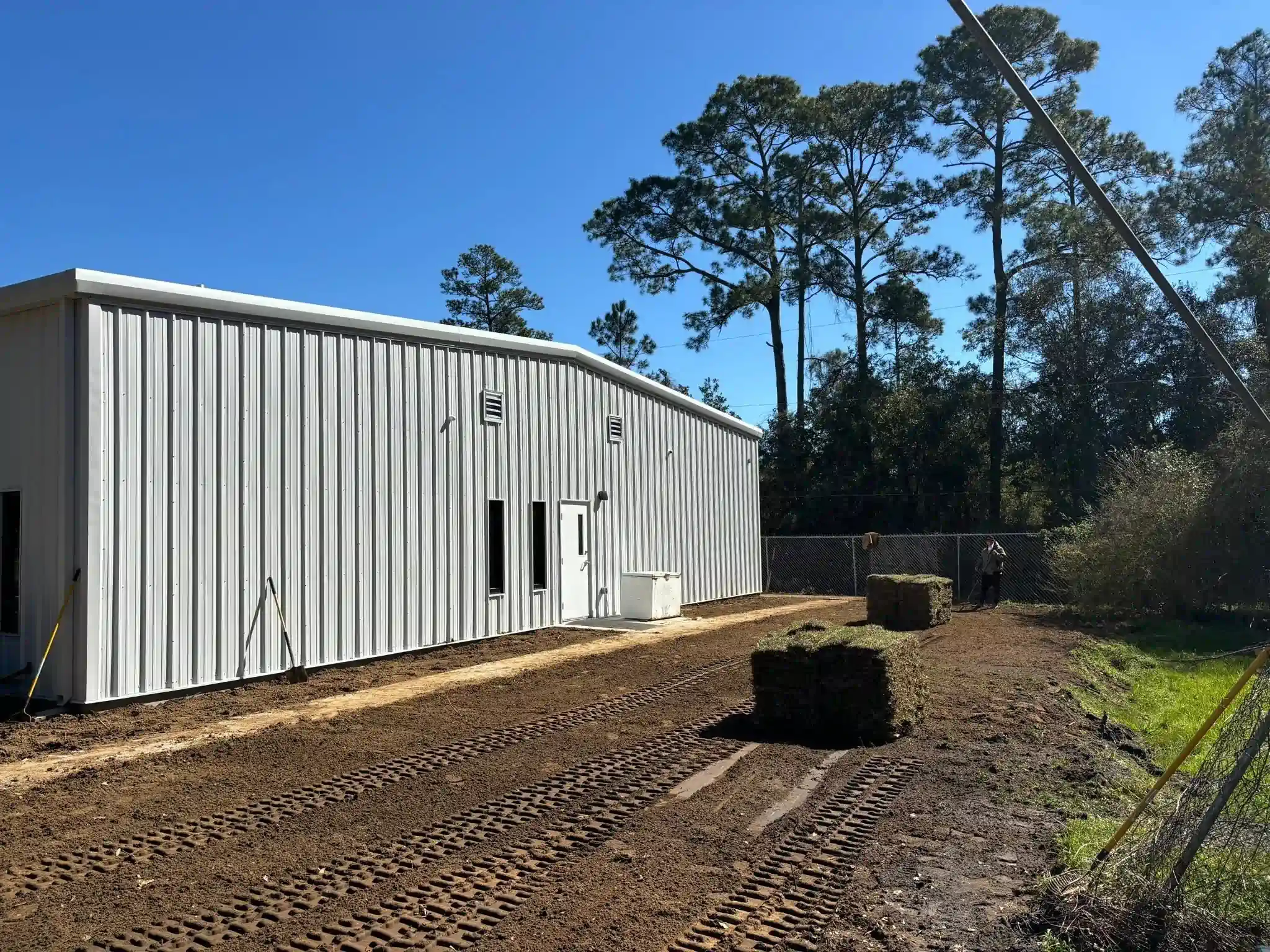 Exterior view of a white metal industrial building, with a muddy dirt path and three rolls of turf grass in front.