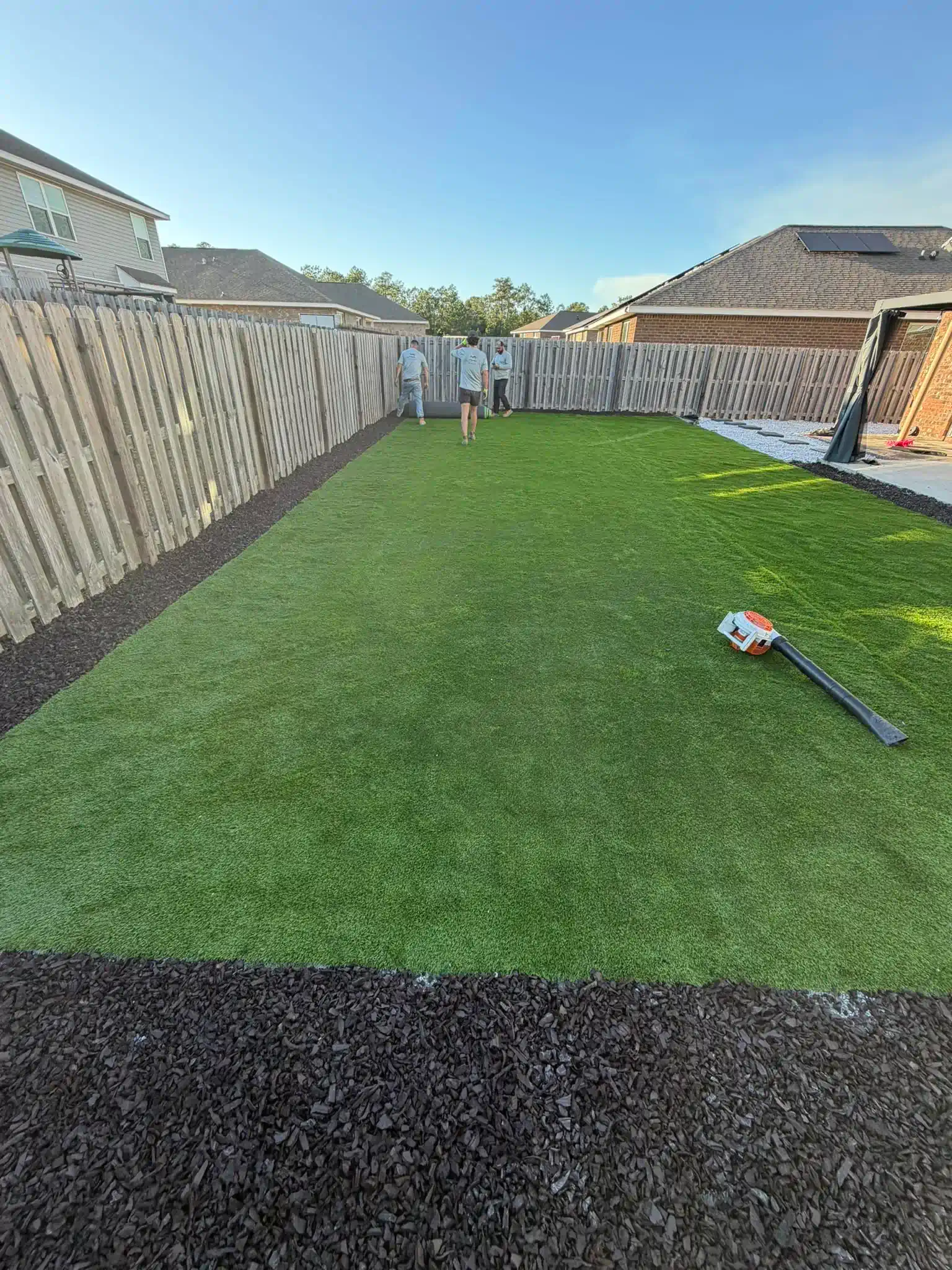 Backyard with green artificial grass, fenced in, person standing, and landscaping.