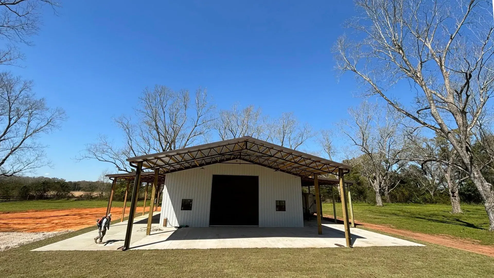 White barn with a dark door under a metal roof, wooden posts. Blue sky, sparse trees.