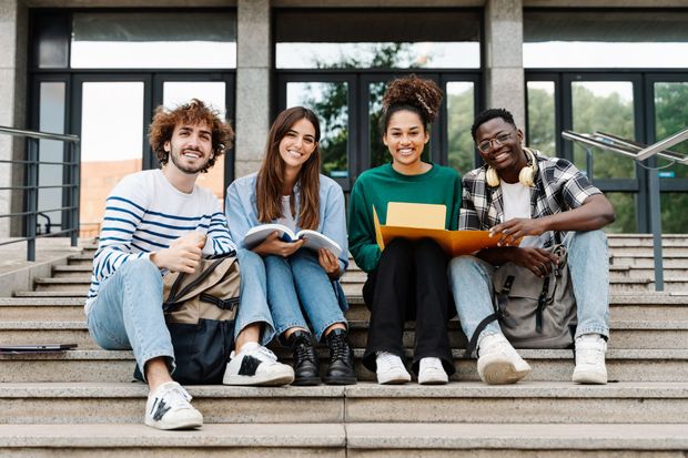 Four students smiling while sitting on university building steps, holding books and a folder.