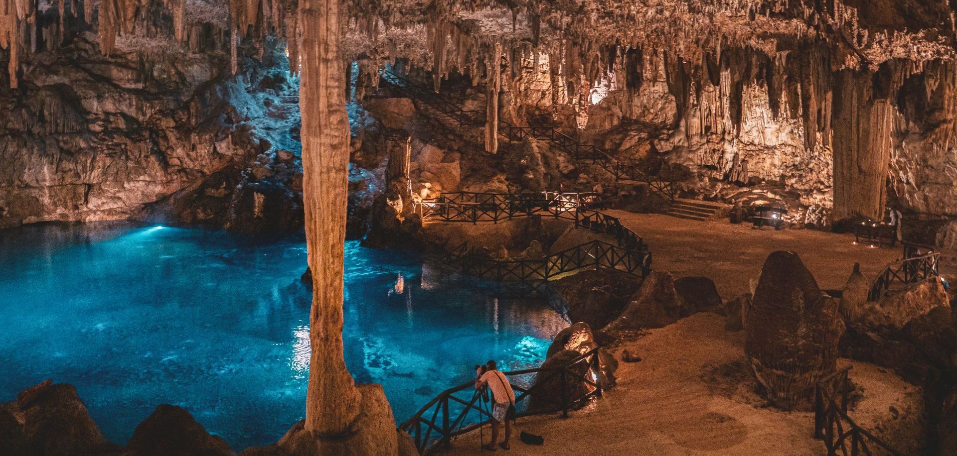 A man is standing on a bridge in a cave with a pool of water.