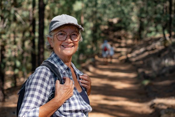 An elderly woman is hiking in the woods with a backpack.