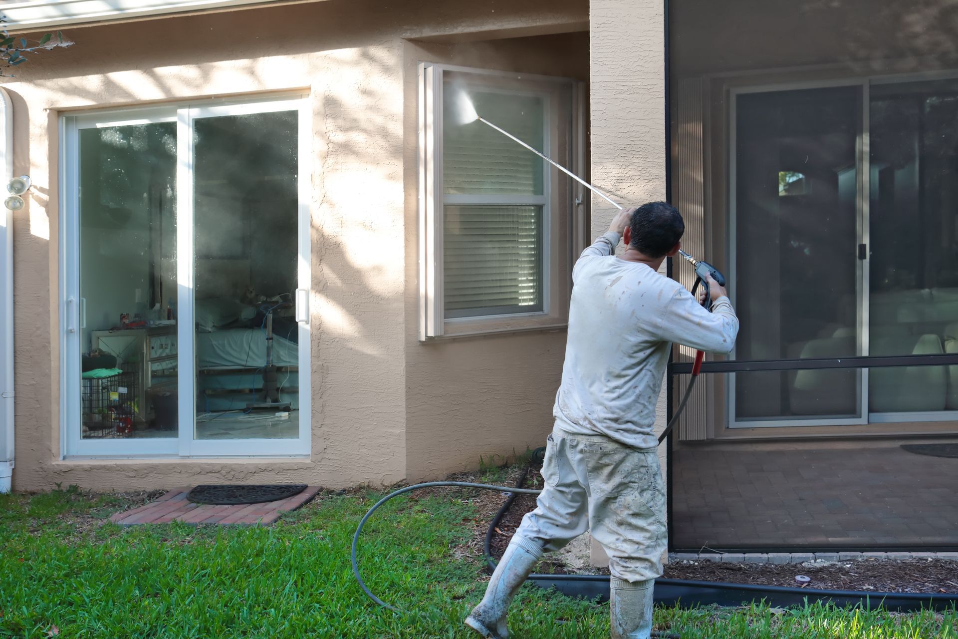 Man pressure washing the exterior of a house with sliding glass doors and windows.