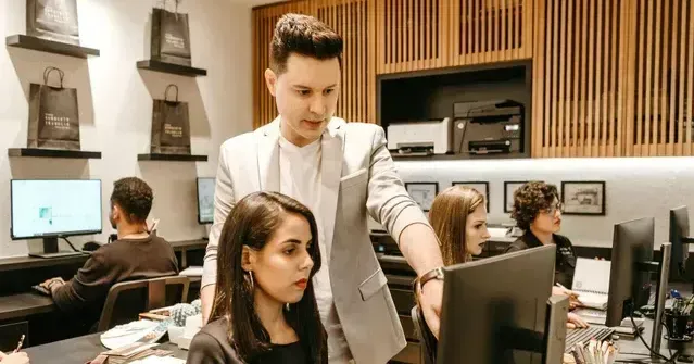 A man is standing next to a woman in front of a computer in an office.