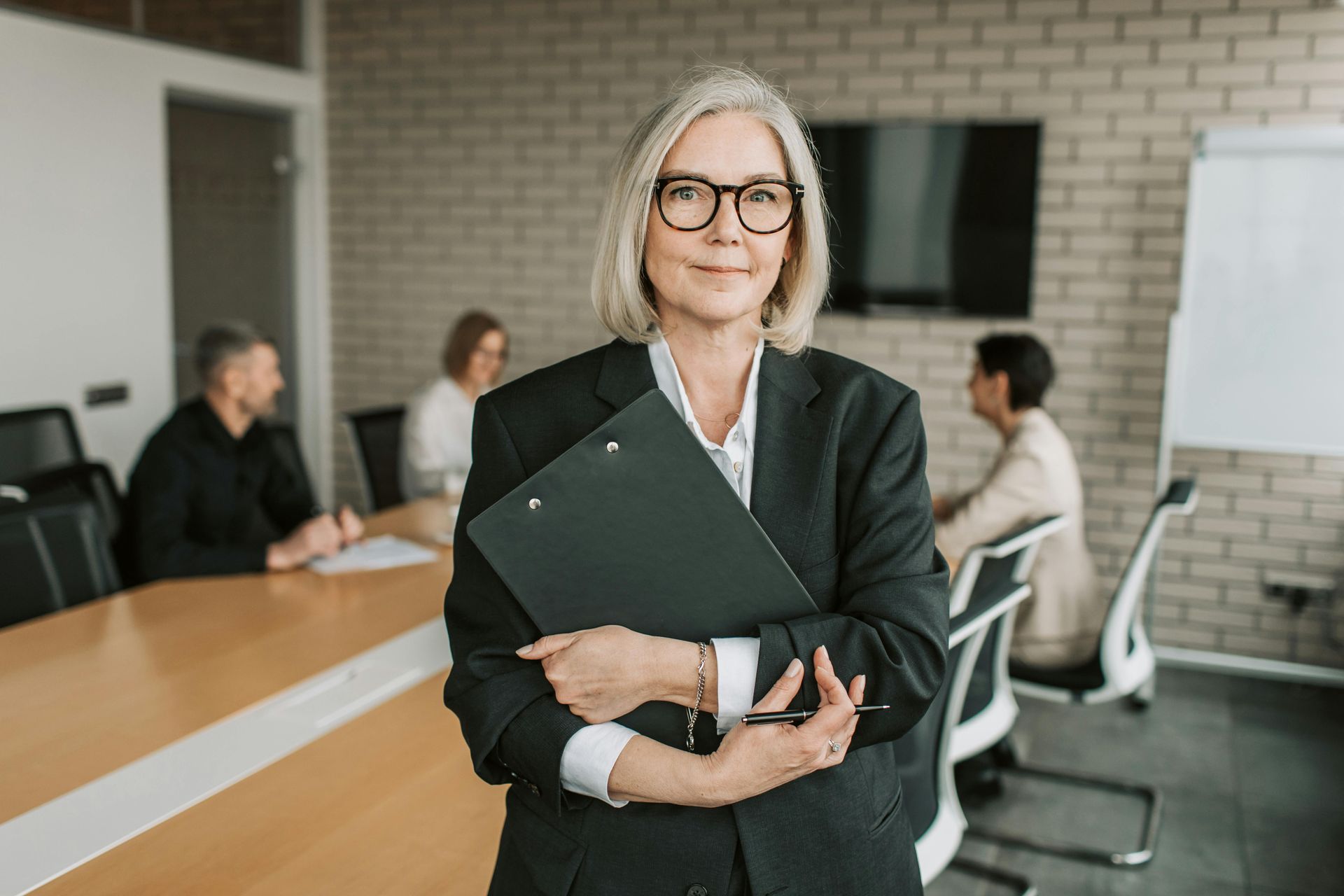 A woman in a suit and glasses is holding a clipboard in a conference room.