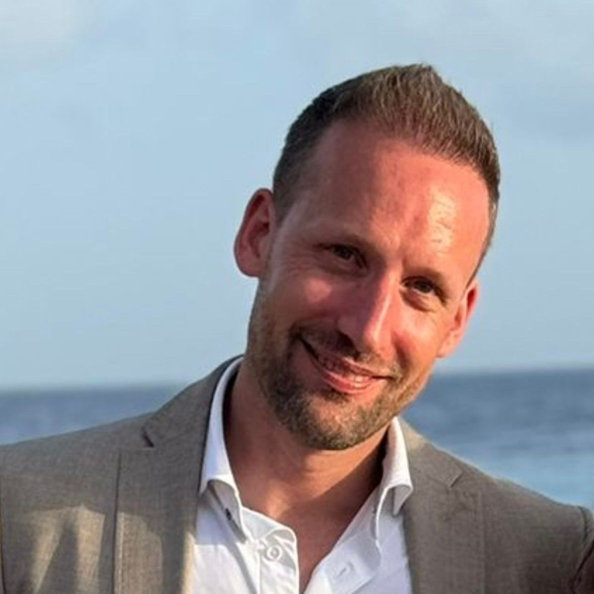 Man in een grijze blazer en wit overhemd, lachend, met de oceaan op de achtergrond.