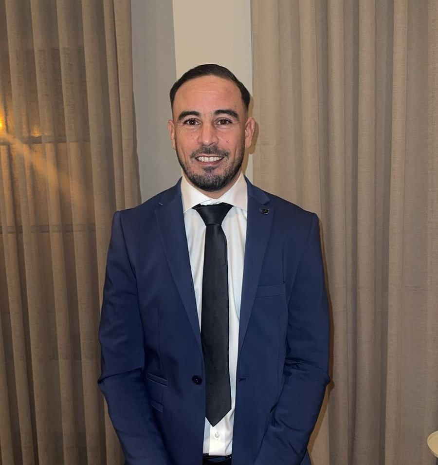 Een man in een blauw pak met zwarte stropdas, glimlachend. Hij staat voor gordijnen.