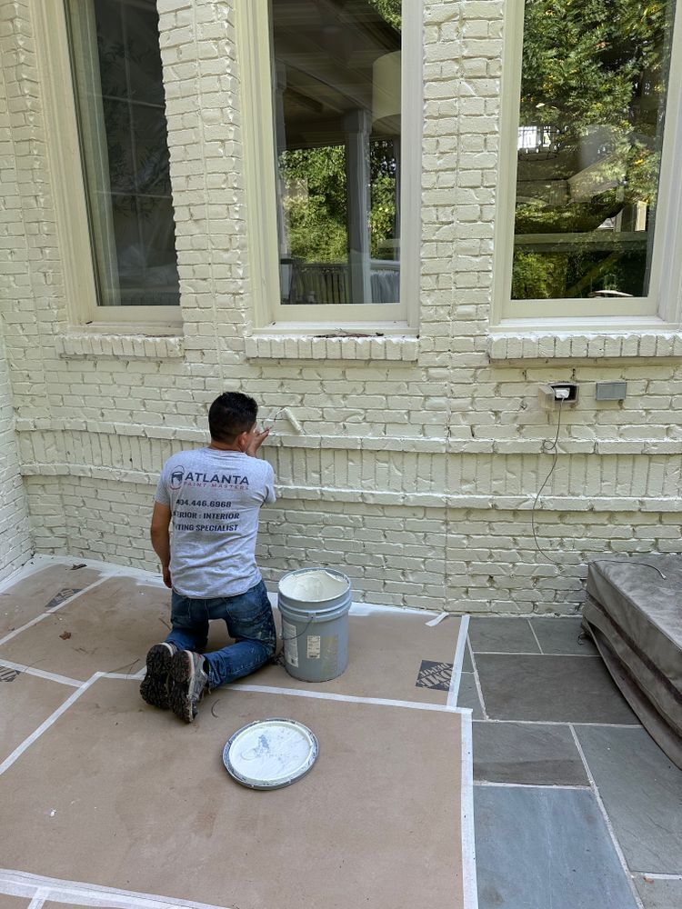 A man is kneeling down while painting a white brick wall.
