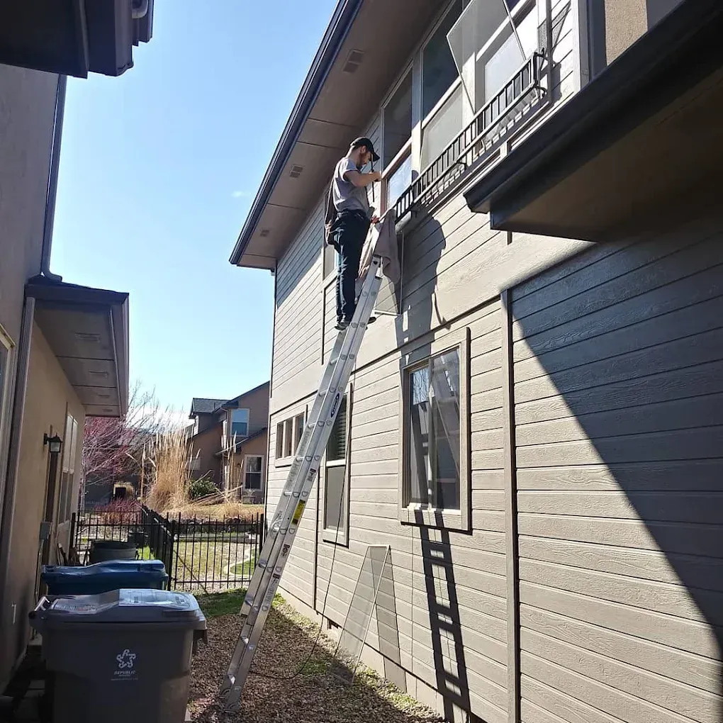 Person on ladder, working on a building's exterior. Sunny day; tan siding, trash cans, and another building are visible.