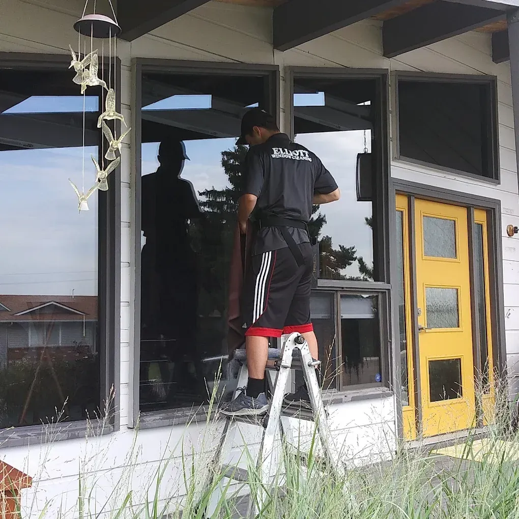 Man on a ladder washing a large window of a house with a yellow door.