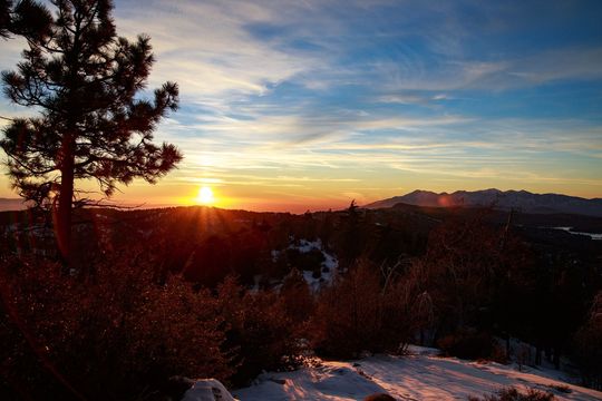 Sunset over a landscape with snow, trees, and mountains, with a bright sun in the center.