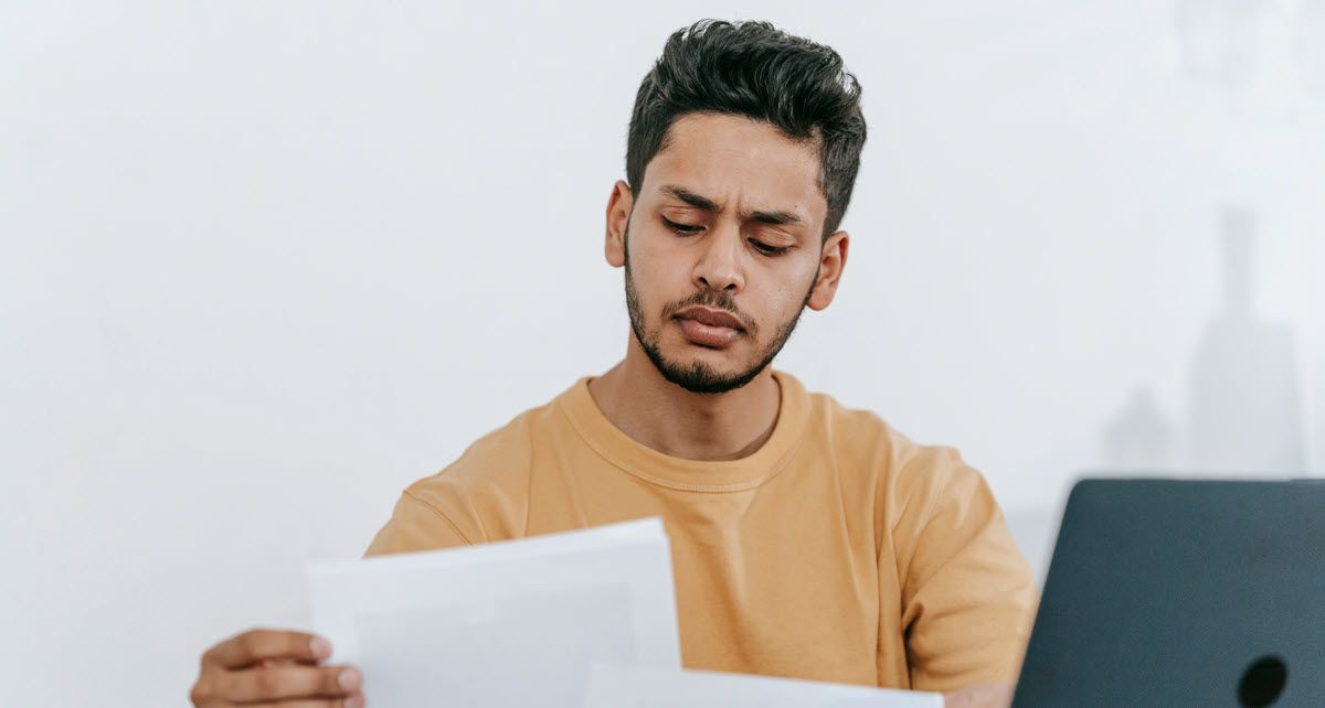 Man in a tan shirt reading a document beside a laptop, looking concerned