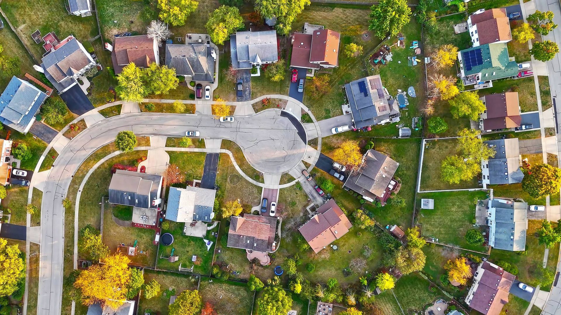 Aerial view of a suburban neighborhood with houses, roads, and a circular drive. Green lawns and colorful trees.