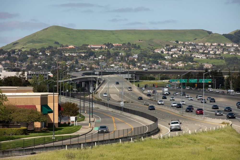 Highway with cars, grassy hills, and residential area under a blue sky.